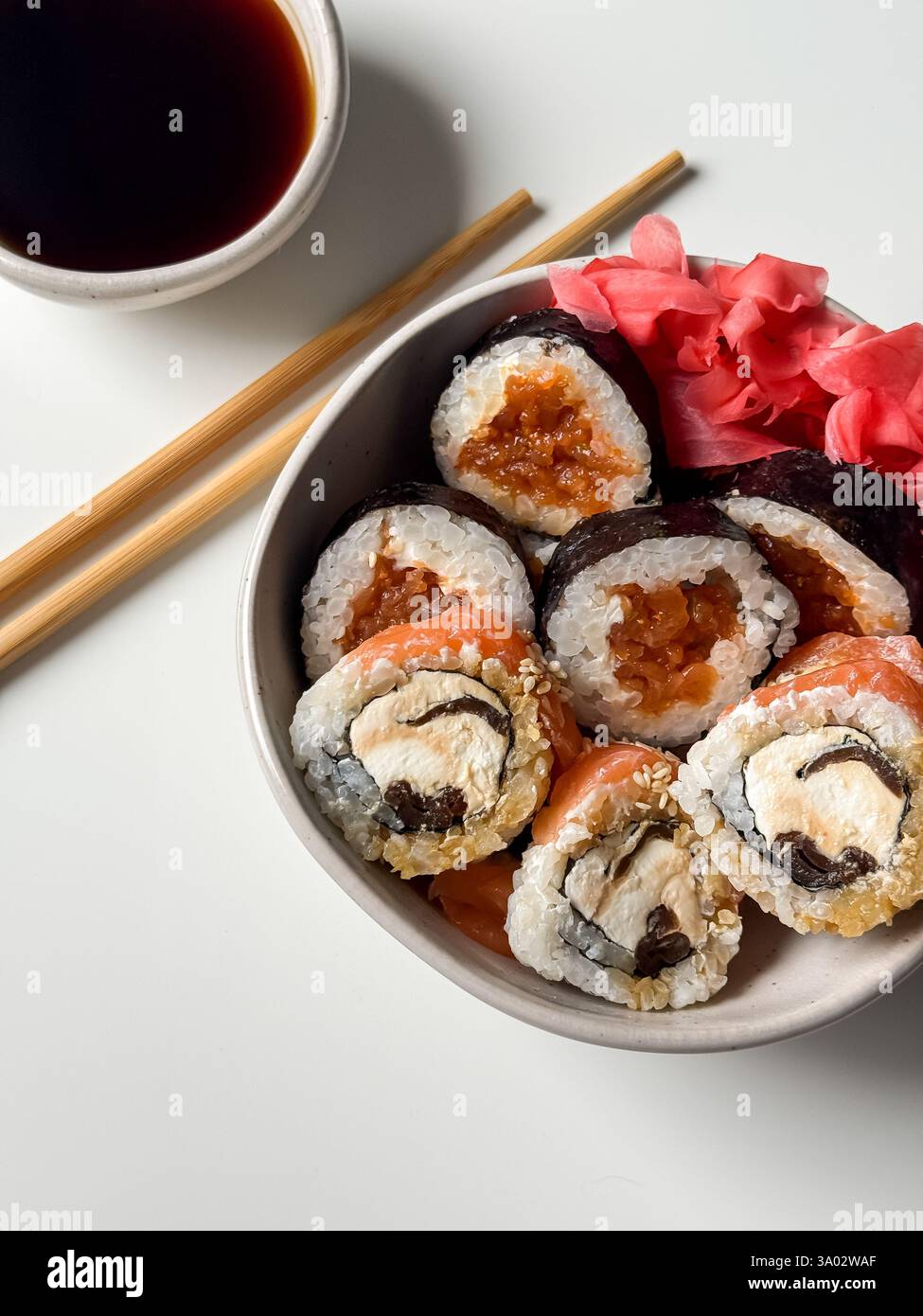 Sushi rollt in einer Schüssel mit rosa Ingwer und Sojasauce und Holzstäbchen auf weißem Tisch Stockfoto