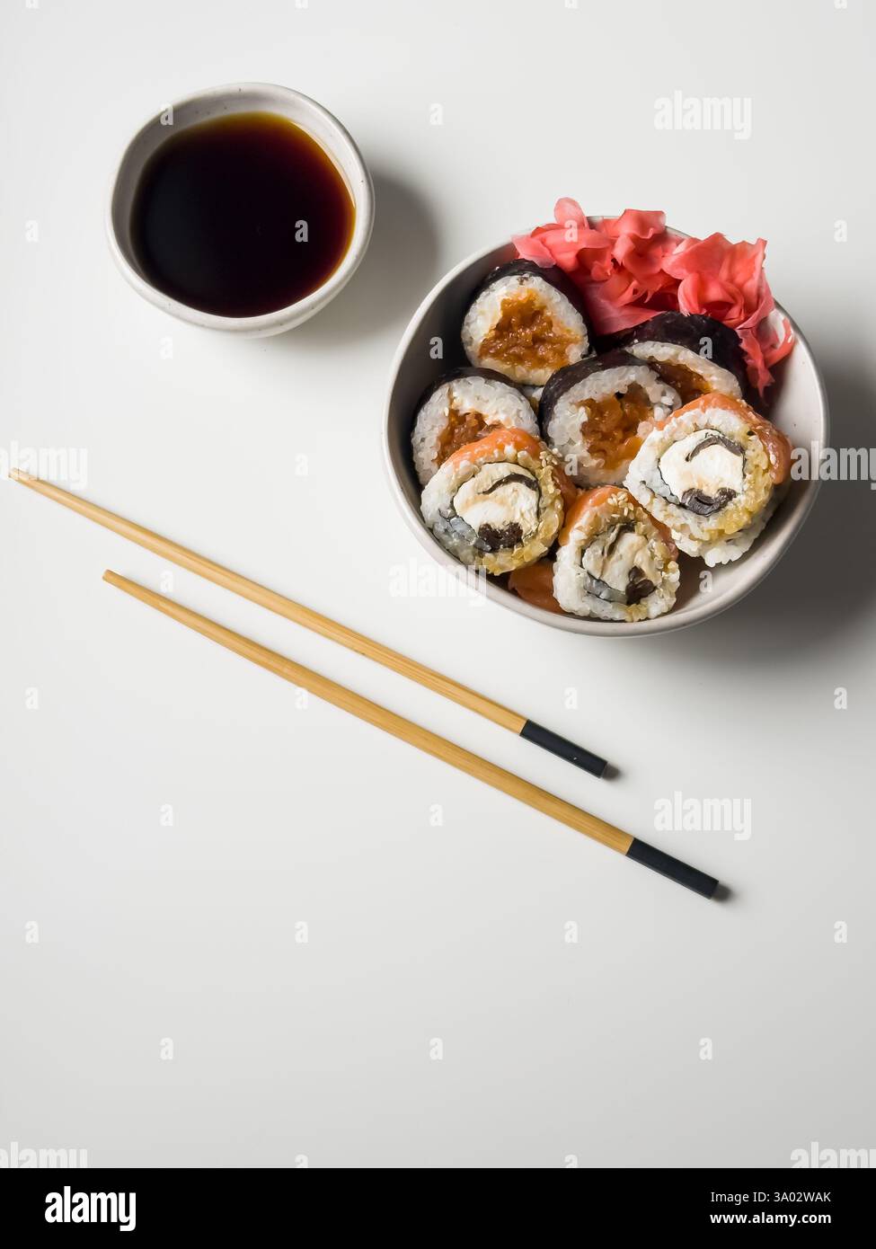Sushi-Brötchen in einer Schüssel mit rosa Ingwer- und Sojasauce und Holzstäbchen auf weißem Tisch Stockfoto