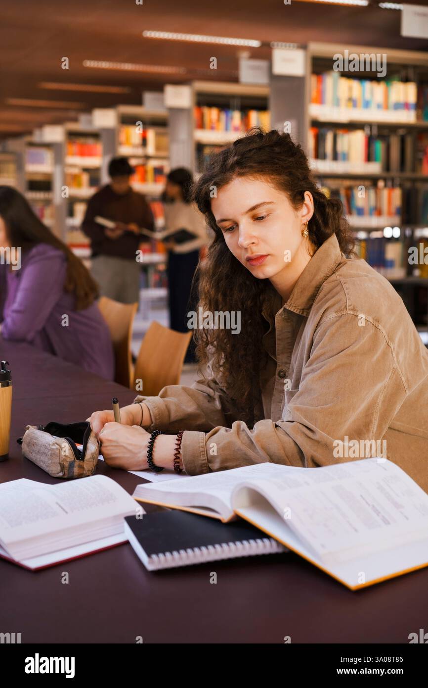 Junge, selbstbewusste Studentin, die im Buch schreibt, während sie am Bücherpult sitzt Stockfoto