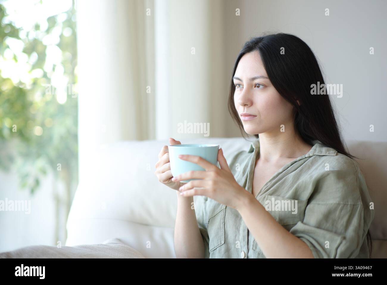Ernsthafte asiatische Frau trinkt Kaffee oder Tee und denkt darüber nach, zu Hause auf einer Couch zu sitzen Stockfoto