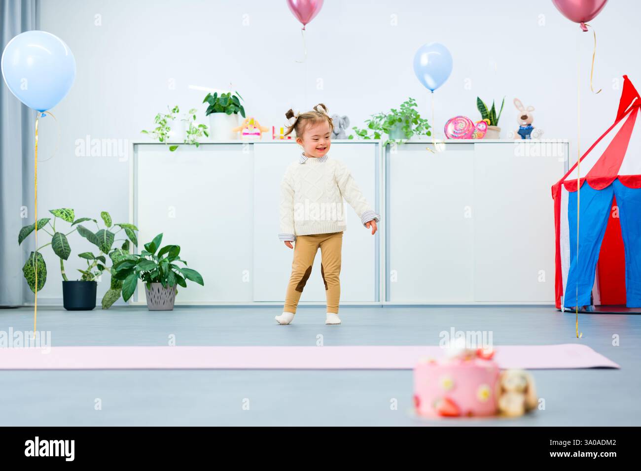 Niedliches 3-jähriges Mädchen mit Down-Syndrom feiert ihren Geburtstag in einem mit Ballons dekorierten Spielzimmer. Kindergeburtstag. Stockfoto