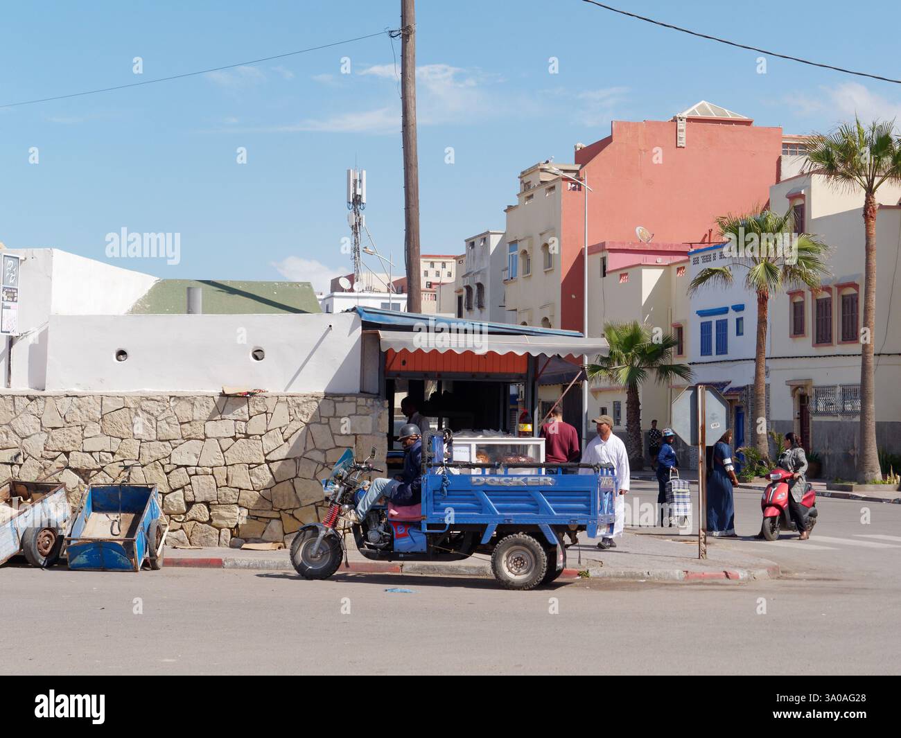 Mann mit Motorrad und Anhänger neben Wagen wartet an der Straßenecke, während sich andere Leute in Essaouira, Marokko, versammeln. März 2025 Stockfoto