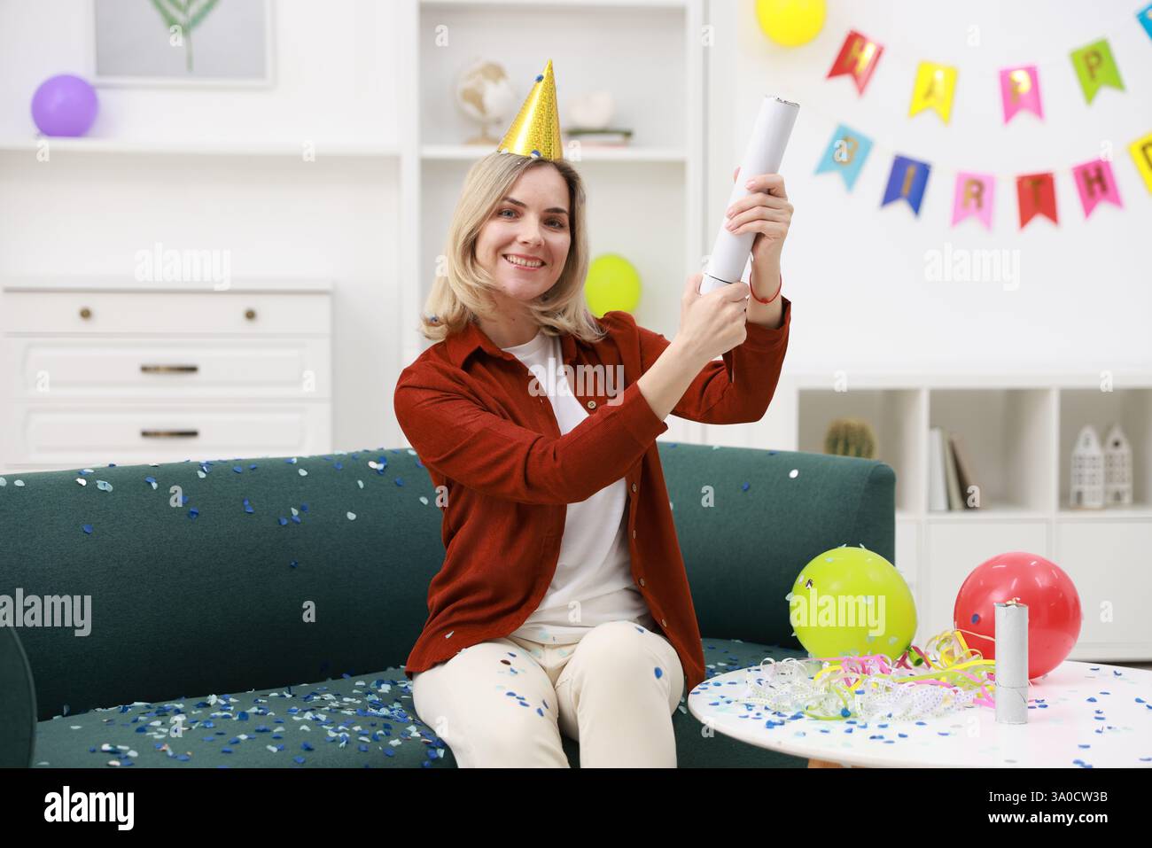 Glückliche Frau, die Konfetti-Popper auf dem Sofa in das Zimmer sprengt, das für eine Party eingerichtet ist Stockfoto