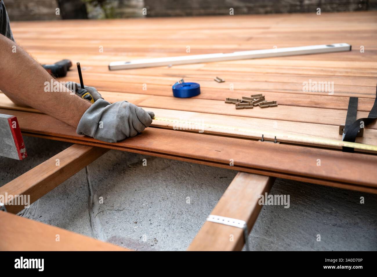 Ein Arbeiter, der Handschuhe trägt, misst und markiert eine Holzdiele beim Bau eines Holzdecks, wobei Werkzeuge und Befestigungselemente verstreut sind. Stockfoto