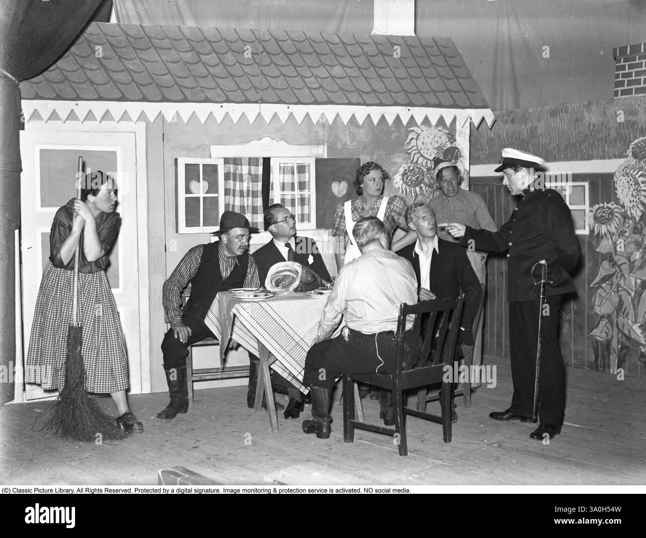 Theater im Jahr 1940. Die Schauspieler des Stücks Jolanta werden in ihren Kostümen auf der Bühne während der Rehersal gesehen. Eine lebendige Szene auf einem Bühnenbild. Die Kulisse zeigt ein malerisches, rustikales Haus mit einem geneigten Dach, karierten Vorhängen in den Fenstern und einer Tür, die an ein Landhaus erinnert. Die Kulisse bietet skurrile, handgezeichnete Sonnenblumen und andere dekorative Elemente, die einen verspielten, theatralischen Touch verleihen. Das Foto spiegelt den Humor und den Charme einer schwedischen Theaterproduktion aus der Mitte des 20. Jahrhunderts wider, in der die Darsteller ihre Rollen in einer unbeschwerten, ländlichen oder häuslichen Umgebung verkörpern. Stockfoto