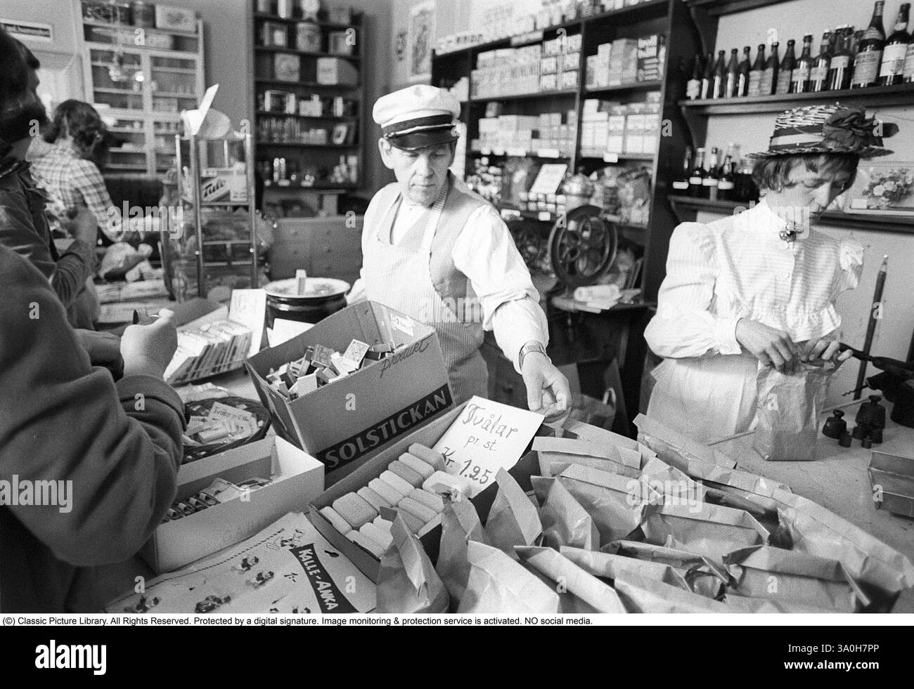 Ein Gemischtwarenmuseum in den 1970er Jahren Fotos, die in einem Gemischtwarenladen aufgenommen wurden und das Erscheinungsbild von vor 100 Jahren festhalten, werden für die Besucher ausgestellt, die dort einkaufen. Die Bilder zeigen eine nostalgische Szene, in der Kunden Waren über den Tresen kaufen, serviert von einem dahinter stehenden Ladenbesitzer, der Waren auf traditionelle, praktische Weise verkauft. Die Einrichtung erinnert an eine vergangene Ära des Handels und verbindet Geschichte mit der Gegenwart, da moderne Käufer denselben Raum erleben, der einst in den Jahrhunderte alten Schnappschüssen dargestellt wurde. Kristoffersson Ref. 8-22 Stockfoto