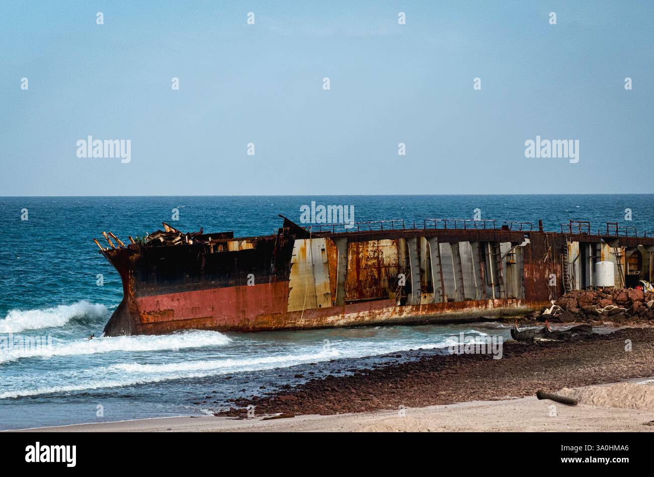 Ein großes, rostiges Schiffswrack liegt an den Ufern von Socotra, das teilweise im kristallklaren Wasser untergetaucht ist, mit einem leuchtend blauen Himmel darüber. Stockfoto