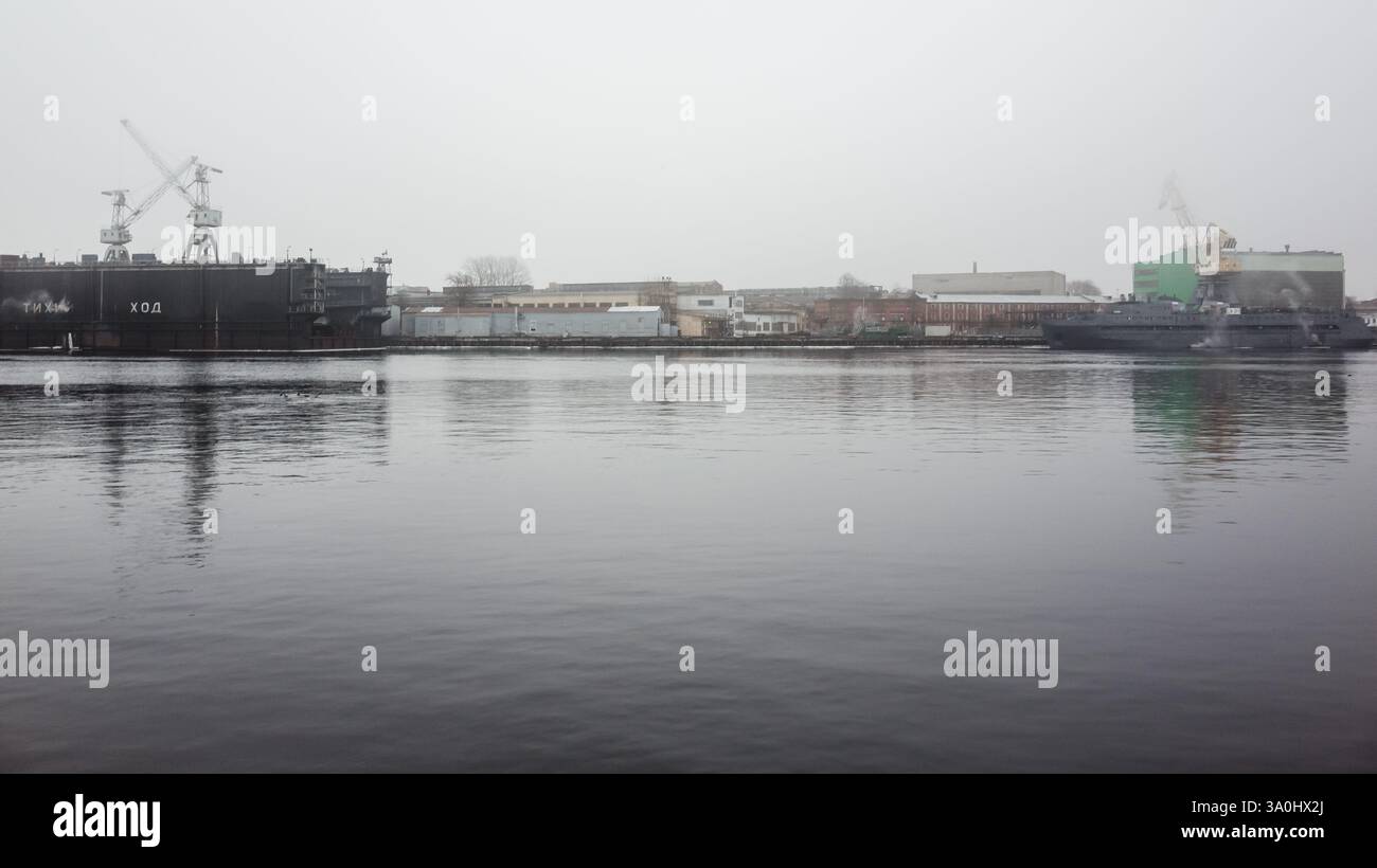 Ein ruhiger Blick auf einen Hafen mit Industrieschiffen, Bauwerken und Docks, umgeben von leichtem Nebel. Küste der Newa, St. Petersburg, Russland Stockfoto