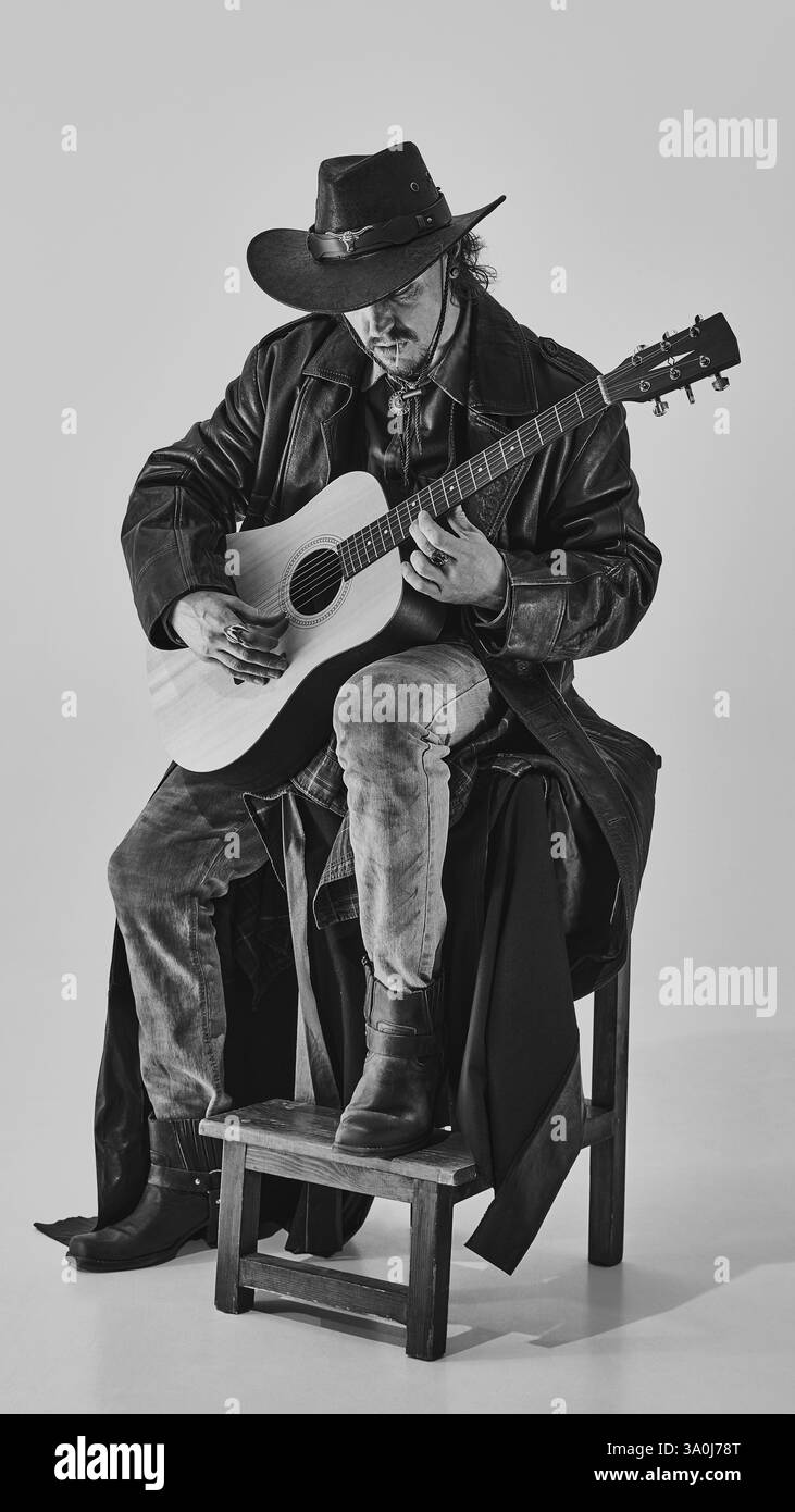 Schwarz-weißes Porträt eines Cowboys, der auf Holzhocker sitzt, mit tiefer Konzentration Gitarre spielt, einen dunklen Ledermantel und Hut trägt Stockfoto