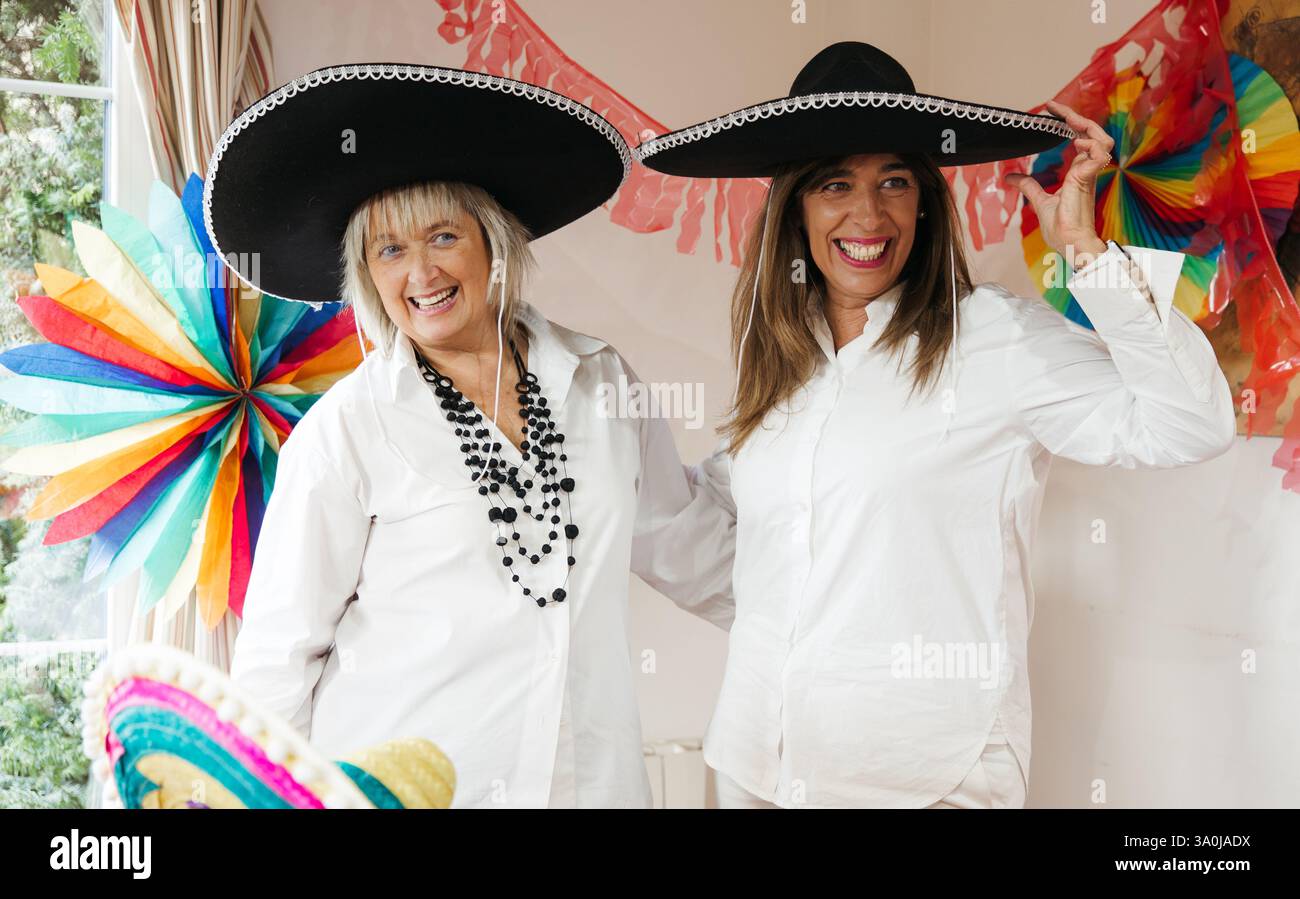 Zwei fröhliche Seniorinnen in weiß gekleidet und tragen schwarze Mariachi Sombreros, die ein festliches Ereignis mit farbenfrohen Dekorationen feiern. Stockfoto
