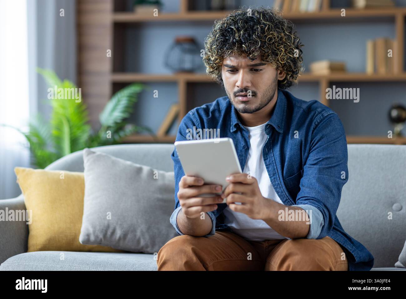 Ein junger Mann mit lockigen Haaren sieht verwirrt aus, während er in einem modernen Wohnzimmer auf seinem Tablet stöbert. Er scheint von etwas überrascht zu sein, das er sieht. Stockfoto