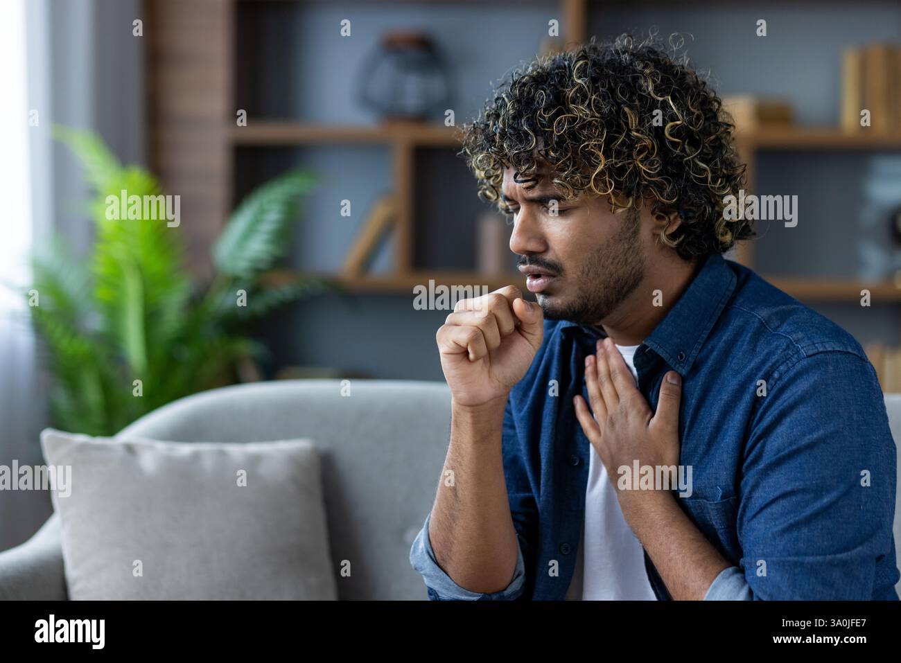 Mann mit lockigem Haar Husten, während er auf einem Sofa in einer inneren Umgebung sitzt, möglicherweise mit Symptomen einer Krankheit oder Allergie. Stockfoto