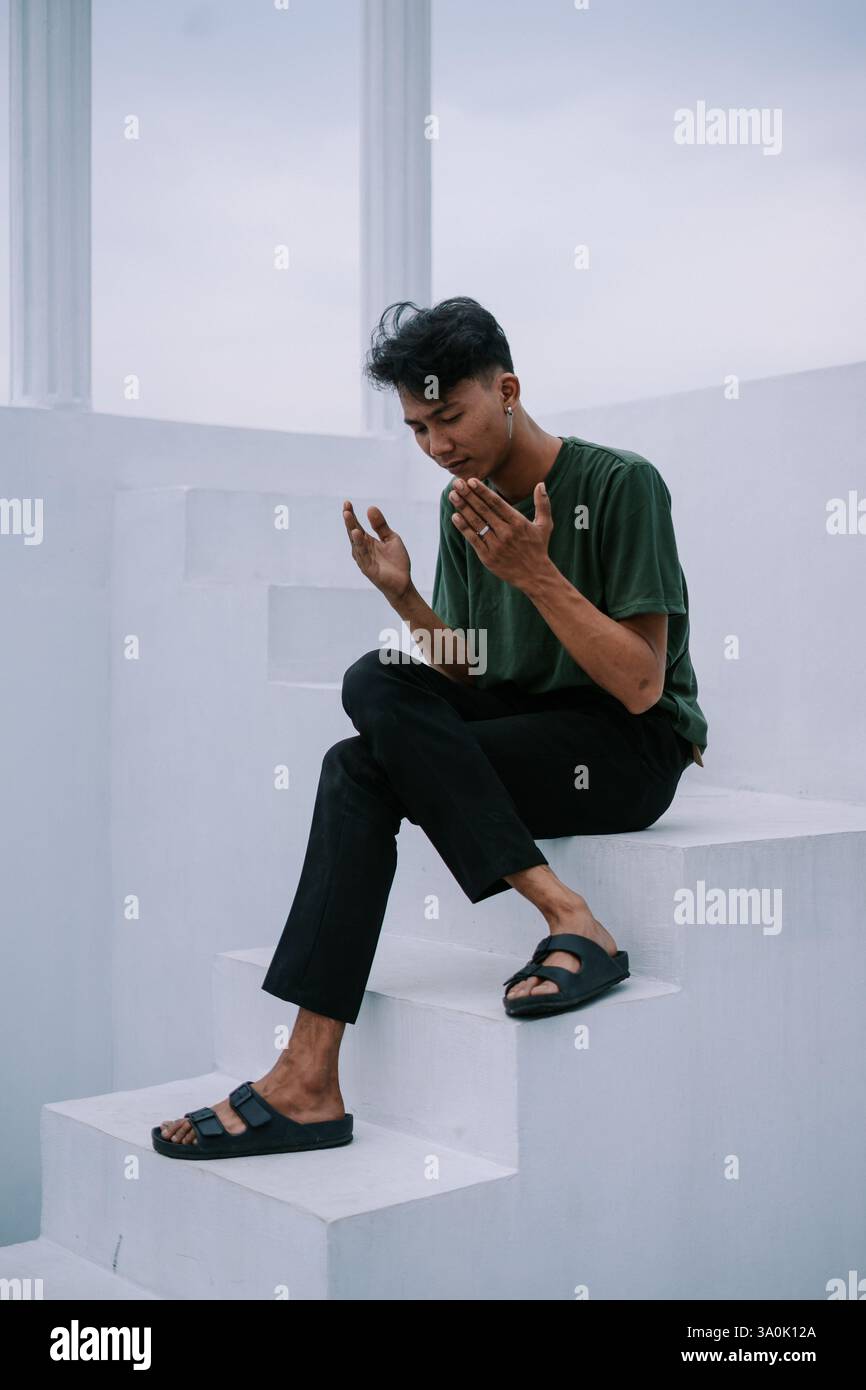 Ein junger Mann sitzt auf einer minimalistischen weißen Treppe, verloren in Gedanken. Sein kontemplativer Ausdruck deutet auf Selbstbetrachtung und Reflexion über den Lebensweg hin. Ca Stockfoto