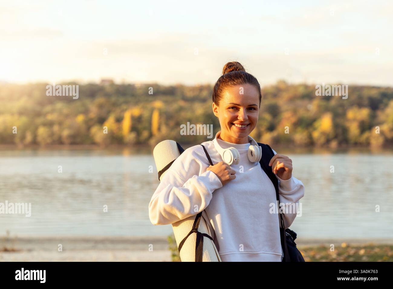 Fitte und aktive Frau mit aufgerollter Yogamatte, die sich nach dem Workout draußen am Fluss entspannt Stockfoto