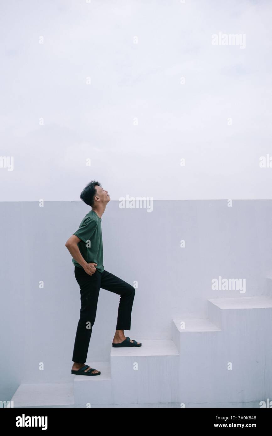 Ein junger Mann klettert selbstbewusst die minimalistische weiße Treppe hinauf und blickt nach oben. Das Bild erinnert an Themen wie Ehrgeiz, Fortschritt und Blick in die Zukunft wi Stockfoto