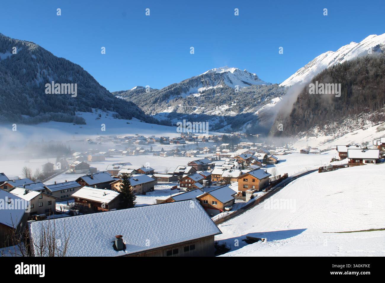 Winterliches Alpenpanorama in Au, Österreich – Schneebedeckte Berge, nebelverhangene Wälder und ein idyllisches Bergdorf im Sonnenschein. Stockfoto