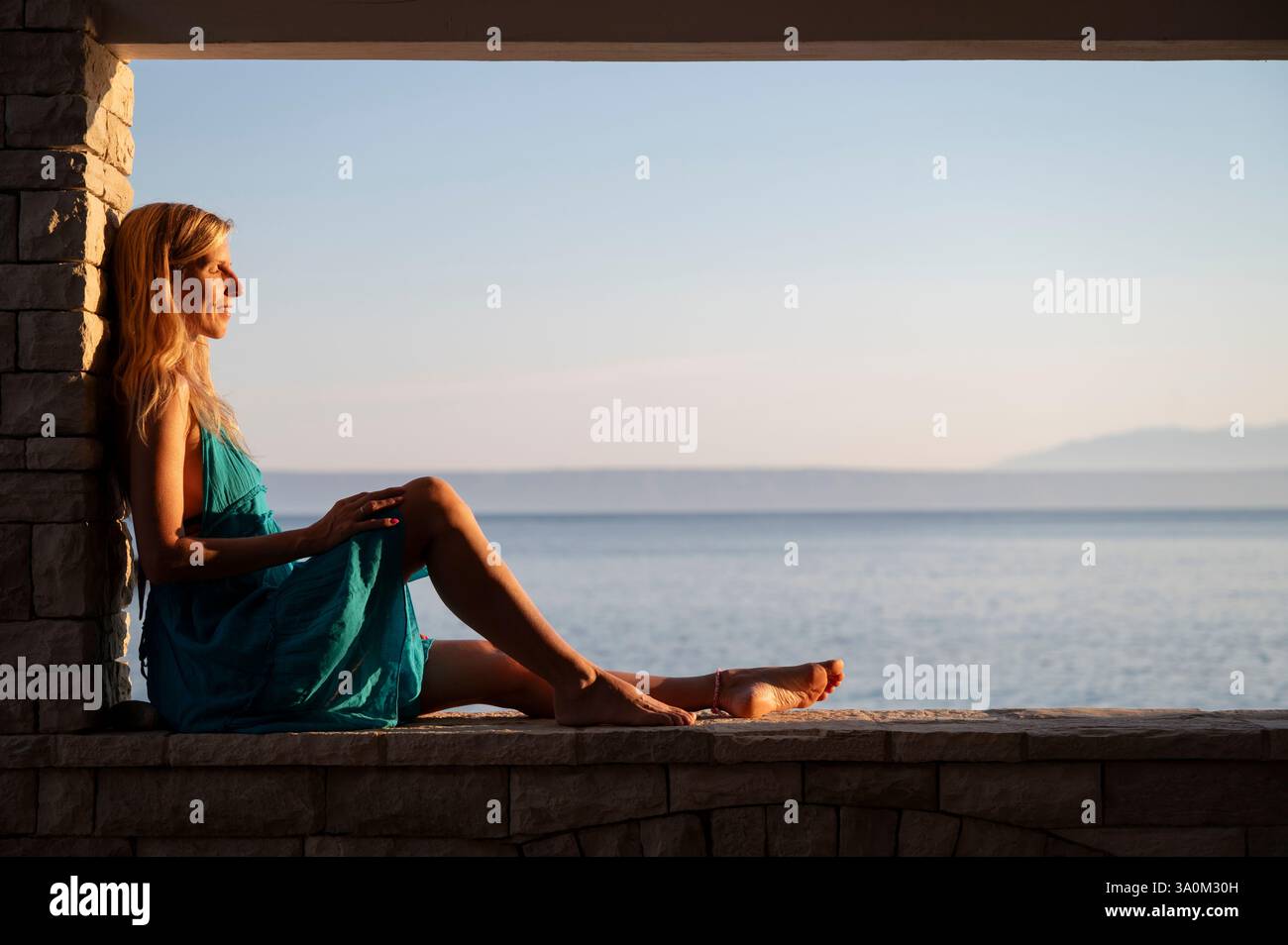 Eine Frau in türkisfarbenem Kleid entspannt sich am Meer, in warmes goldenes Licht getaucht, und blickt auf das ruhige Meer mit fernen Bergen am Horizont. Stockfoto