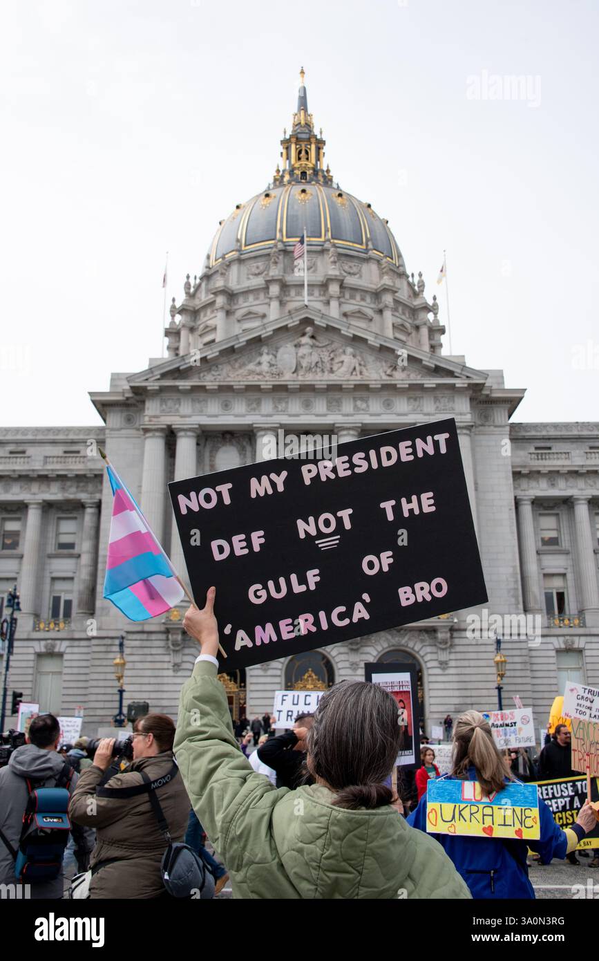 San Francisco, USA. März 2025. Demonstranten versammeln sich vor dem Rathaus von San Francisco, um gegen die Befehle von Präsident Trump und gegen die Aktionen von Elon Musk und seinem DOGENTEAM zu protestieren. Ein Demonstrant hält eine Trans-Flagge und ein Schild mit der Aufschrift "nicht mein Präsident, def nicht der Golf von Amerika", in Bezug auf Trumps kürzliche Umbenennung des Golfs von Mexiko in den Golf von Amerika. Quelle: Shelly Rivoli/Alamy Live News Stockfoto