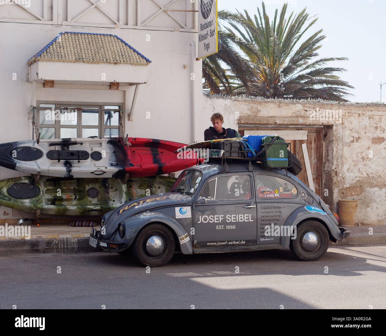 Man packt einen alten grauen VW-Käfer mit Benzinkanister auf einem Dachträger neben einem Kayaks-Grundstück in Essaouira. März 2025 Stockfoto