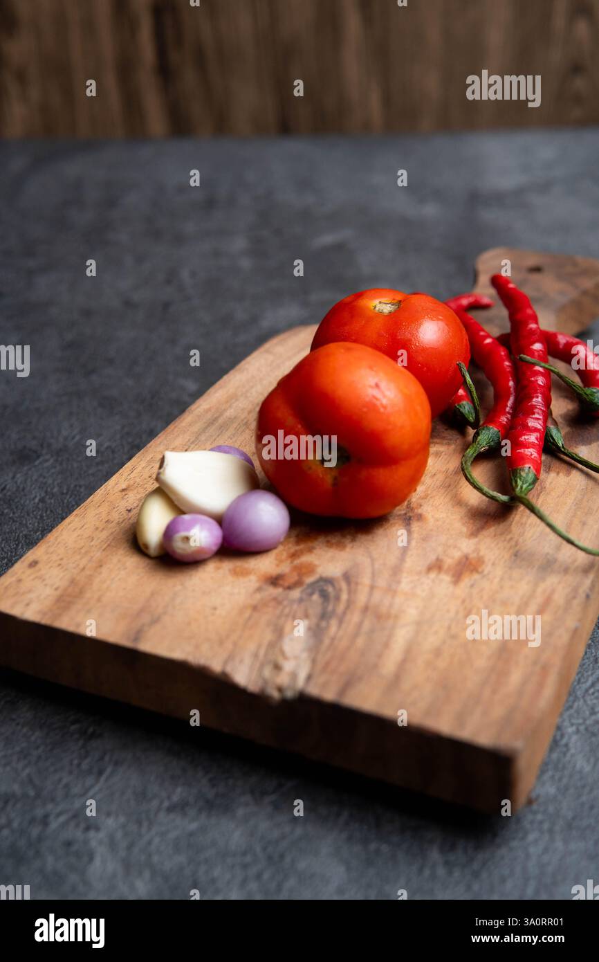 Ein hölzernes Schneidebrett mit frischen Zutaten, darunter reife rote Tomaten, rote Chilischoten, Schalotten und Knoblauch. Stockfoto