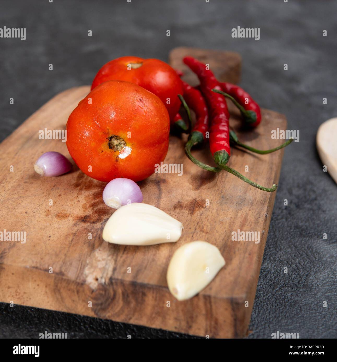 Ein hölzernes Schneidebrett mit frischen Zutaten, darunter reife rote Tomaten, rote Chilischoten, Schalotten und Knoblauch. Stockfoto