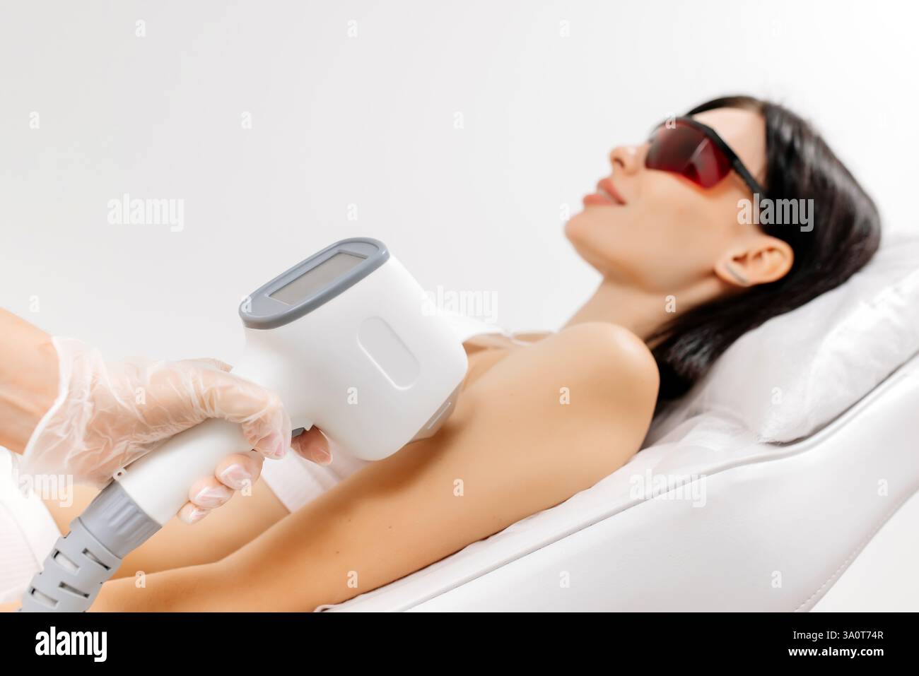 Körperpflegekonzept. Laserhaarentfernung. Junge weiße Frau in Schutzbrille liegt auf dem Sofa, weibliche Kosmetikerin, die Laser-Haarentfernung macht Stockfoto Körperpflegekonzept. Laserhaarentfernung. Junge weiße Frau in Schutzbrille liegt auf dem Sofa, weibliche Kosmetikerin, die Laser-Haarentfernung macht Stockfoto