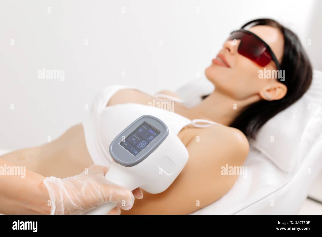 Körperpflegekonzept. Laserhaarentfernung. Junge weiße Frau in Schutzbrille liegt auf dem Sofa, weibliche Kosmetikerin, die Laser-Haarentfernung macht Stockfoto Körperpflegekonzept. Laserhaarentfernung. Junge weiße Frau in Schutzbrille liegt auf dem Sofa, weibliche Kosmetikerin, die Laser-Haarentfernung macht Stockfoto