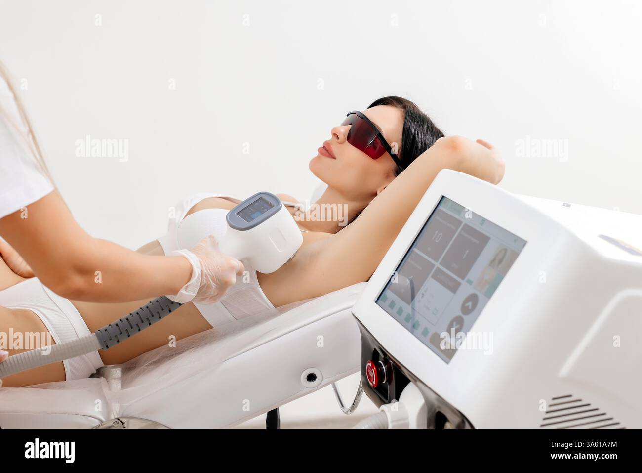 Körperpflegekonzept. Laserhaarentfernung. Junge weiße Frau in Schutzbrille liegt auf dem Sofa, weibliche Kosmetikerin, die Laser-Haarentfernung macht Stockfoto Körperpflegekonzept. Laserhaarentfernung. Junge weiße Frau in Schutzbrille liegt auf dem Sofa, weibliche Kosmetikerin, die Laser-Haarentfernung macht Stockfoto