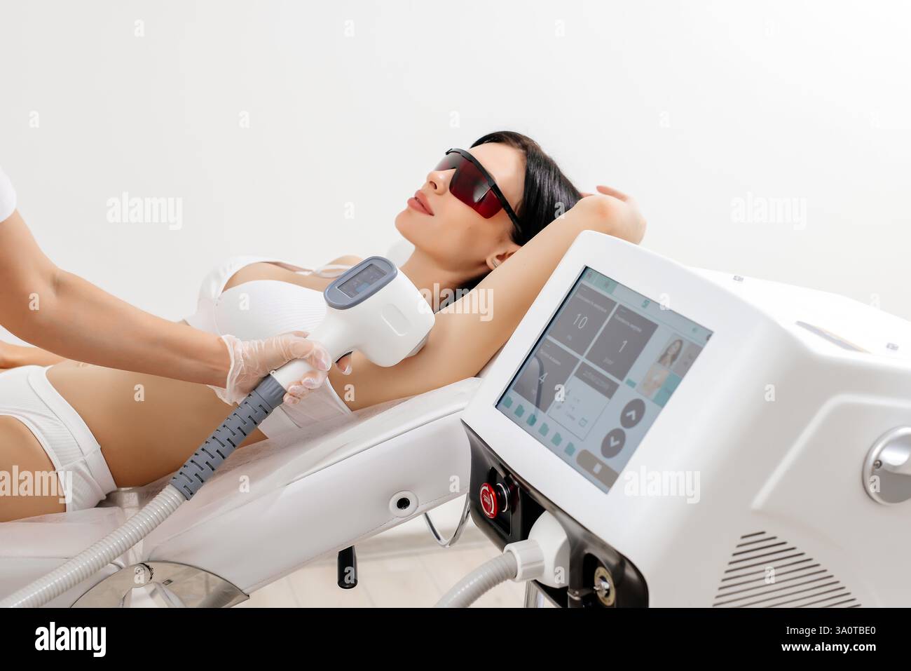 Körperpflegekonzept. Laserhaarentfernung. Junge weiße Frau in Schutzbrille liegt auf dem Sofa, weibliche Kosmetikerin, die Laser-Haarentfernung macht Stockfoto Körperpflegekonzept. Laserhaarentfernung. Junge weiße Frau in Schutzbrille liegt auf dem Sofa, weibliche Kosmetikerin, die Laser-Haarentfernung macht Stockfoto