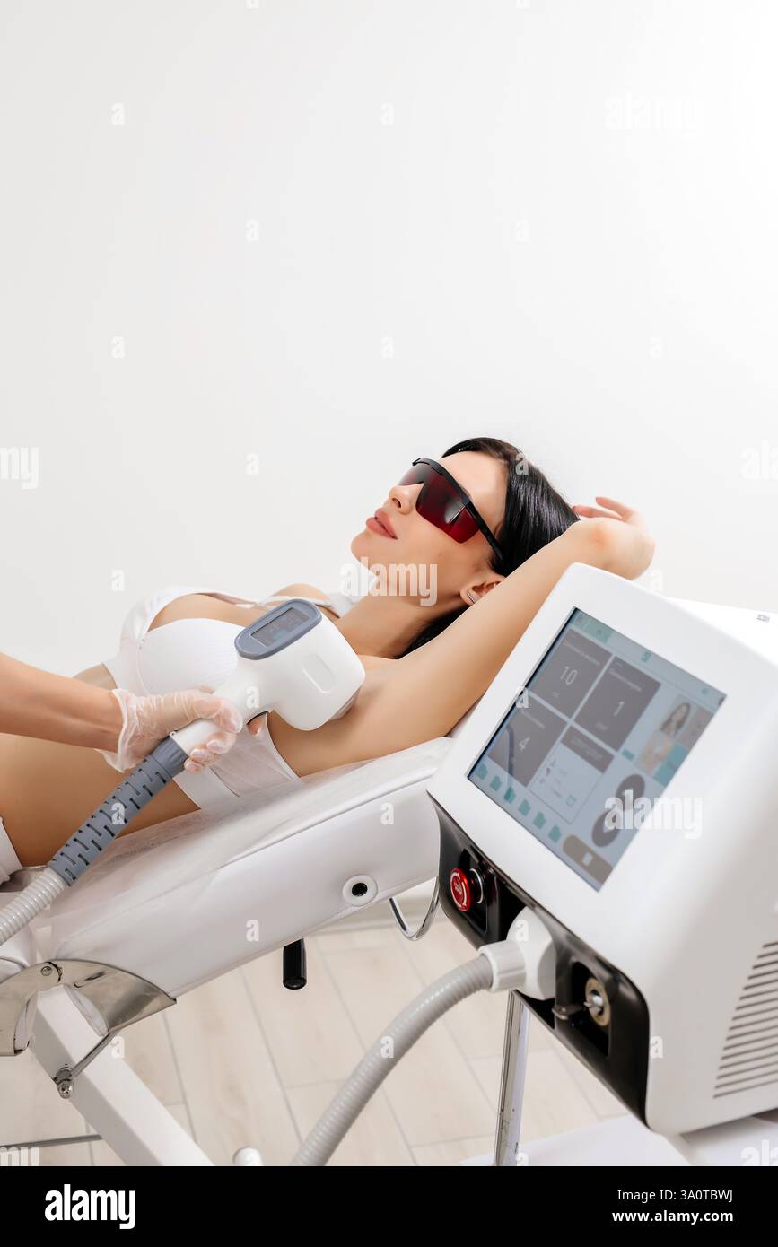 Körperpflegekonzept. Laserhaarentfernung. Junge weiße Frau in Schutzbrille liegt auf dem Sofa, weibliche Kosmetikerin, die Laser-Haarentfernung macht Stockfoto Körperpflegekonzept. Laserhaarentfernung. Junge weiße Frau in Schutzbrille liegt auf dem Sofa, weibliche Kosmetikerin, die Laser-Haarentfernung macht Stockfoto