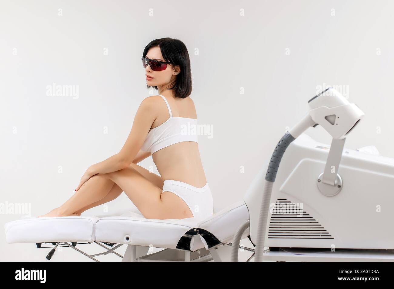 Körperpflegekonzept. Laserhaarentfernung. Junge weiße Frau in Schutzbrille liegt auf dem Sofa, weibliche Kosmetikerin, die Laser-Haarentfernung macht Stockfoto Körperpflegekonzept. Laserhaarentfernung. Junge weiße Frau in Schutzbrille liegt auf dem Sofa, weibliche Kosmetikerin, die Laser-Haarentfernung macht Stockfoto