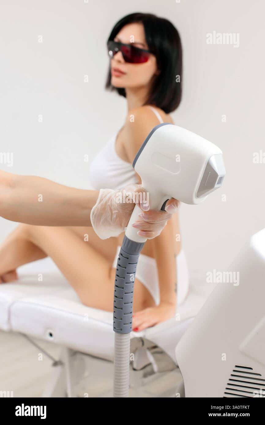 Körperpflegekonzept. Laserhaarentfernung. Junge weiße Frau in Schutzbrille liegt auf dem Sofa, weibliche Kosmetikerin, die Laser-Haarentfernung macht Stockfoto Körperpflegekonzept. Laserhaarentfernung. Junge weiße Frau in Schutzbrille liegt auf dem Sofa, weibliche Kosmetikerin, die Laser-Haarentfernung macht Stockfoto