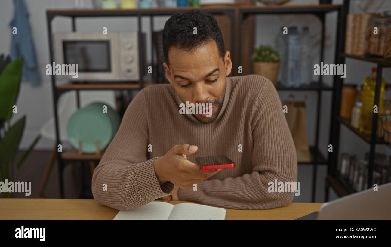 Junger Mann im Büro, der von zu Hause aus mit einem Smartphone arbeitet, umgeben von Büchern und Gadgets, präsentiert eine moderne und dynamische Arbeitsumgebung wi Stockfoto