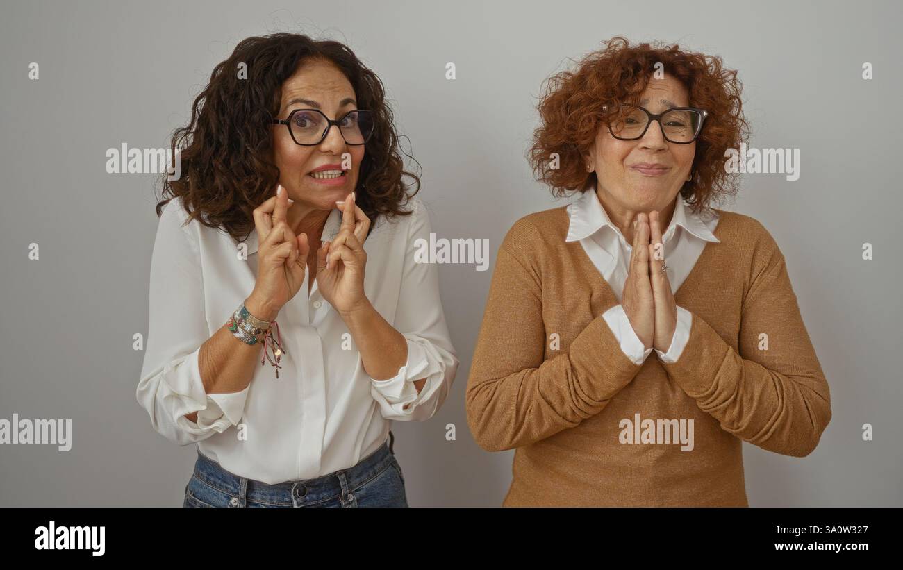 Frauen, die die Finger kreuzen und auf weißem Hintergrund flehen, zeigen eine warme Freundschaft zwischen zwei hispanischen Frauen mittleren Alters, die Hoffnung ausdrücken, zu bekommen Stockfoto