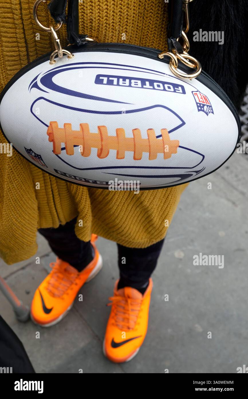 Ältere britische Fußballfan mit NFL Ball Handtasche Mode Tasche Nike Schuhe stehen stolz posiert in einer Londoner Straße England UK 2025 KATHY DEWITT Stockfoto