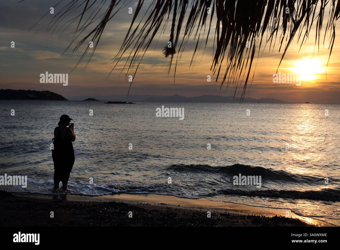 Frau, die im Meer steht und den Sonnenuntergang beobachtet, fotografiert mit Handy Vouliagmeni Beach Attica Griechenland Stockfoto