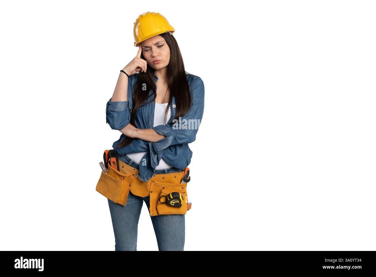 Eine junge Frau mit gelbem Schutzhelm und Denim-Hemd, mit einem Werkzeuggürtel und einem nachdenklichen Ausdruck. Sie reibt sich die Stirn, als ob sie es wäre Stockfoto
