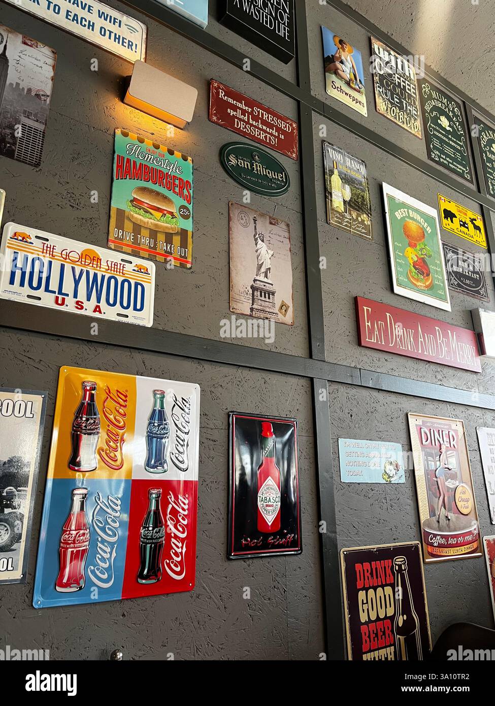 Die Wand eines Cafés ist mit einer Vielzahl von farbenfrohen Logos und Schildern verziert, darunter Coca-Cola-Branding, Hamburger-Bilder und Hollywood-Symbole. - Smartphone-aufgenommenes Stockfoto