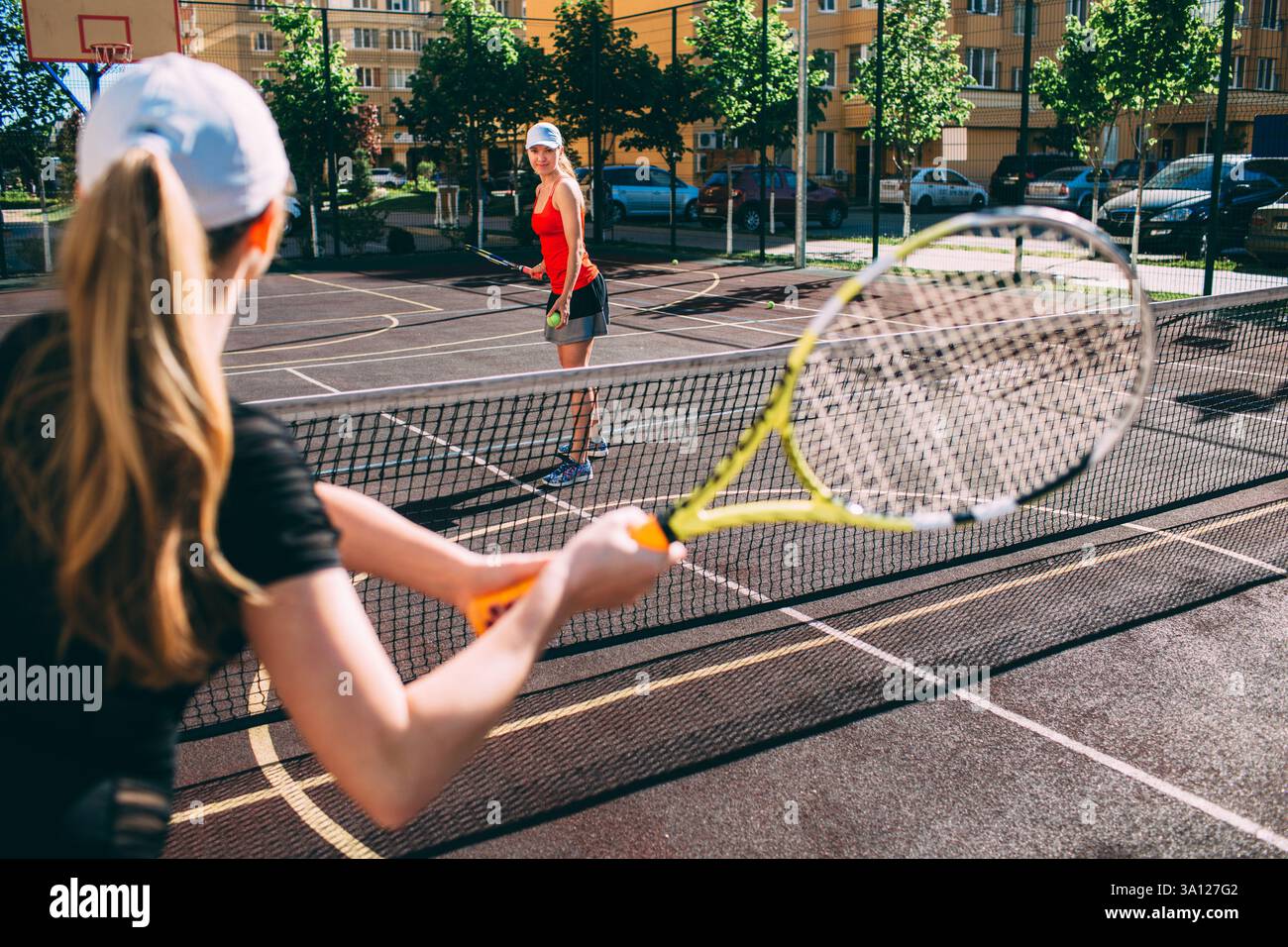 Zwei Einzelpersonen spielen eine freundliche Partie Tennis unter klarem Himmel auf einem von Grünanlagen und Gebäuden umgebenen Außenplatz. Stockfoto