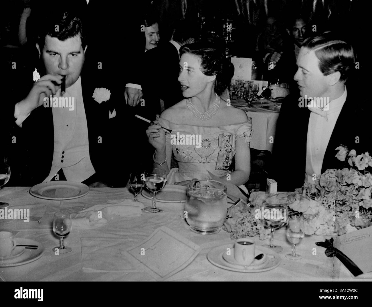 Prinzessin Margaret auf Einem Wohltätigkeitsball -- Prinzessin Margaret, im schulterfreien weißen Satinkleid mit zwei Medaillons ihrer Schwester und ihres Vaters, raucht eine Zigarette in einem langen Halter bei einem Wohltätigkeitsball im Savoy Hotel, London, zur Unterstützung der Dockland Settlement. Mit der Prinzessin rauchen Mr. Jeremy Tree und der Marquess of Blandford. Jeder war beim Tanzen mit der Prinzessin verbunden. Diese alte Phantom-Einsamkeit verfolgte sie. Die Zigarrenrauchende Jeremy Tree und der Marquess of Blandford waren ihre Freunde. Mai 1953. (Foto von Reuterphoto). Stockfoto