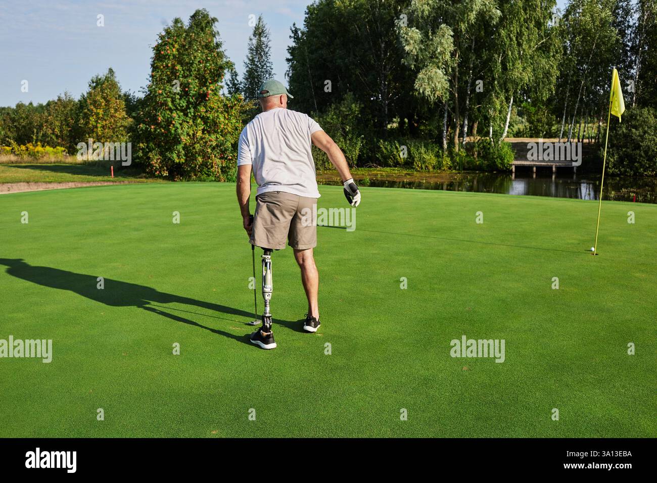Individual mit einer Beinprothese, die sich auf einen lebhaften Golfplatz konzentriert, der von Bäumen umgeben ist, bei Sonnenlicht am Nachmittag. Stockfoto