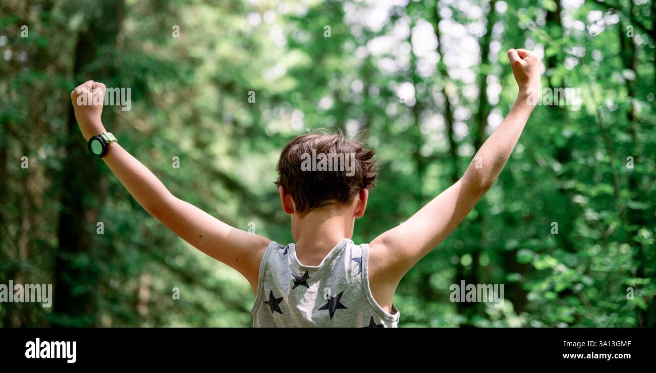 Ein kleiner Junge in einem Tanktop mit Sternmuster hebt triumphierend seine Fäuste in einem üppig grünen Wald und nimmt die Energie der Natur an einem sonnigen Tag auf. Stockfoto