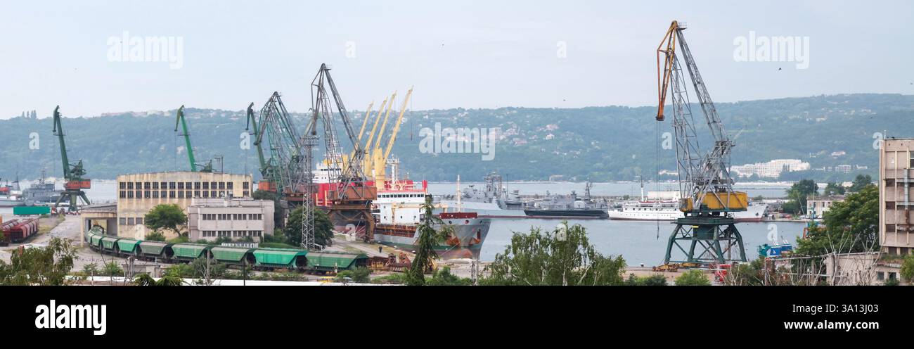 Ein geschäftiger Industriehafen mit Kränen, angedockten Schiffen und Lagerhäusern entlang einer ruhigen Küstenlandschaft unter bedecktem Himmel. Varna, Bulgari Stockfoto
