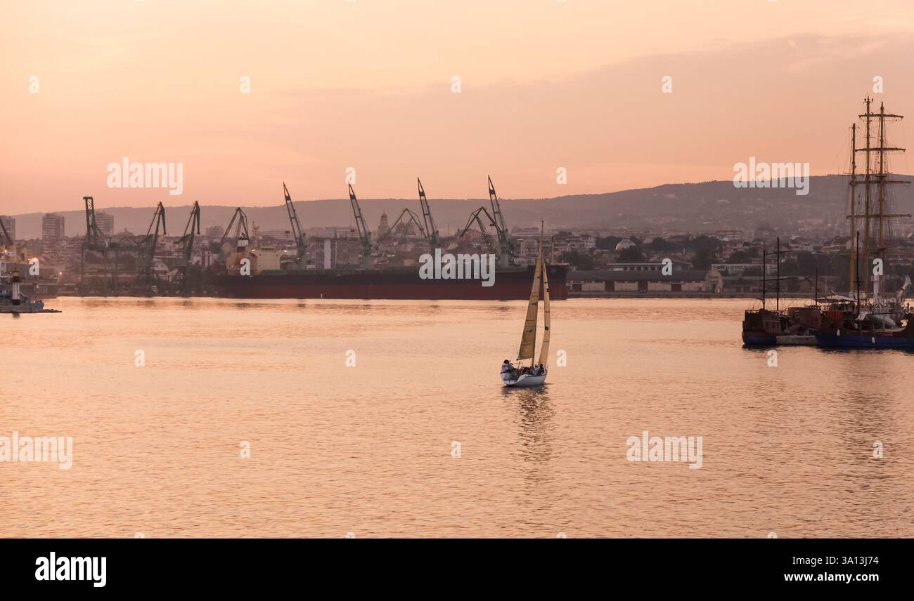 Ruhiger Abendblick mit einem einzigen Segelboot, das auf ruhigen Gewässern vor einer malerischen Skyline fährt, beleuchtet von einem warmen und farbenfrohen Sonnenuntergang, Stockfoto Ruhiger Abendblick mit einem einzigen Segelboot, das auf ruhigen Gewässern vor einer malerischen Skyline fährt, beleuchtet von einem warmen und farbenfrohen Sonnenuntergang, Stockfoto