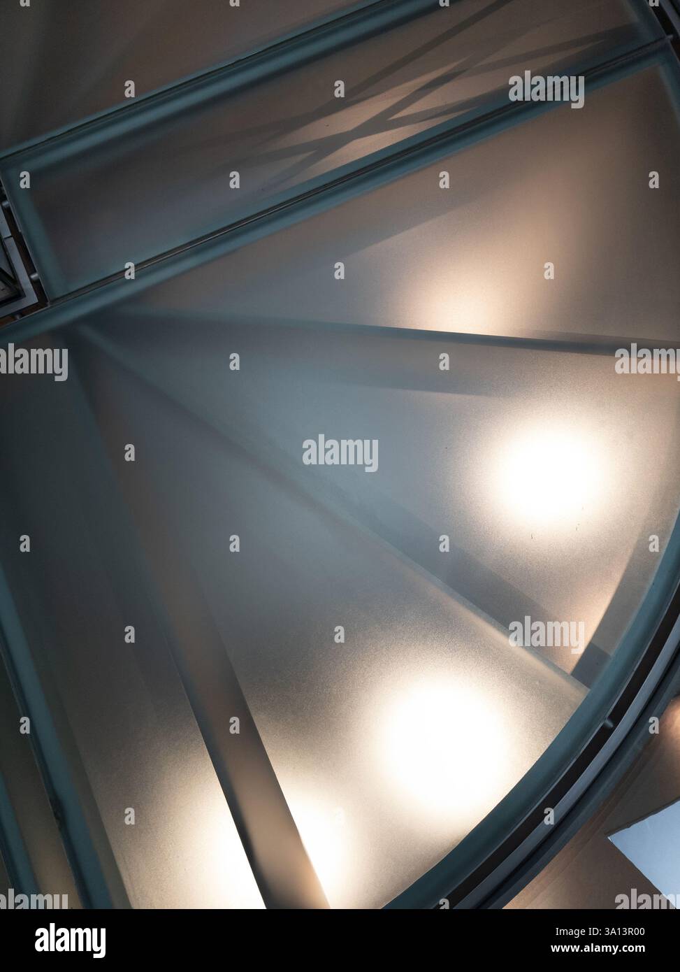 Wendeltreppe mit mattierten Glaspaneelen, die von eleganten Metallschienen eingerahmt werden und von unten sanft beleuchtet werden, unterstreicht die moderne Ästhetik und elegante Architektur Stockfoto