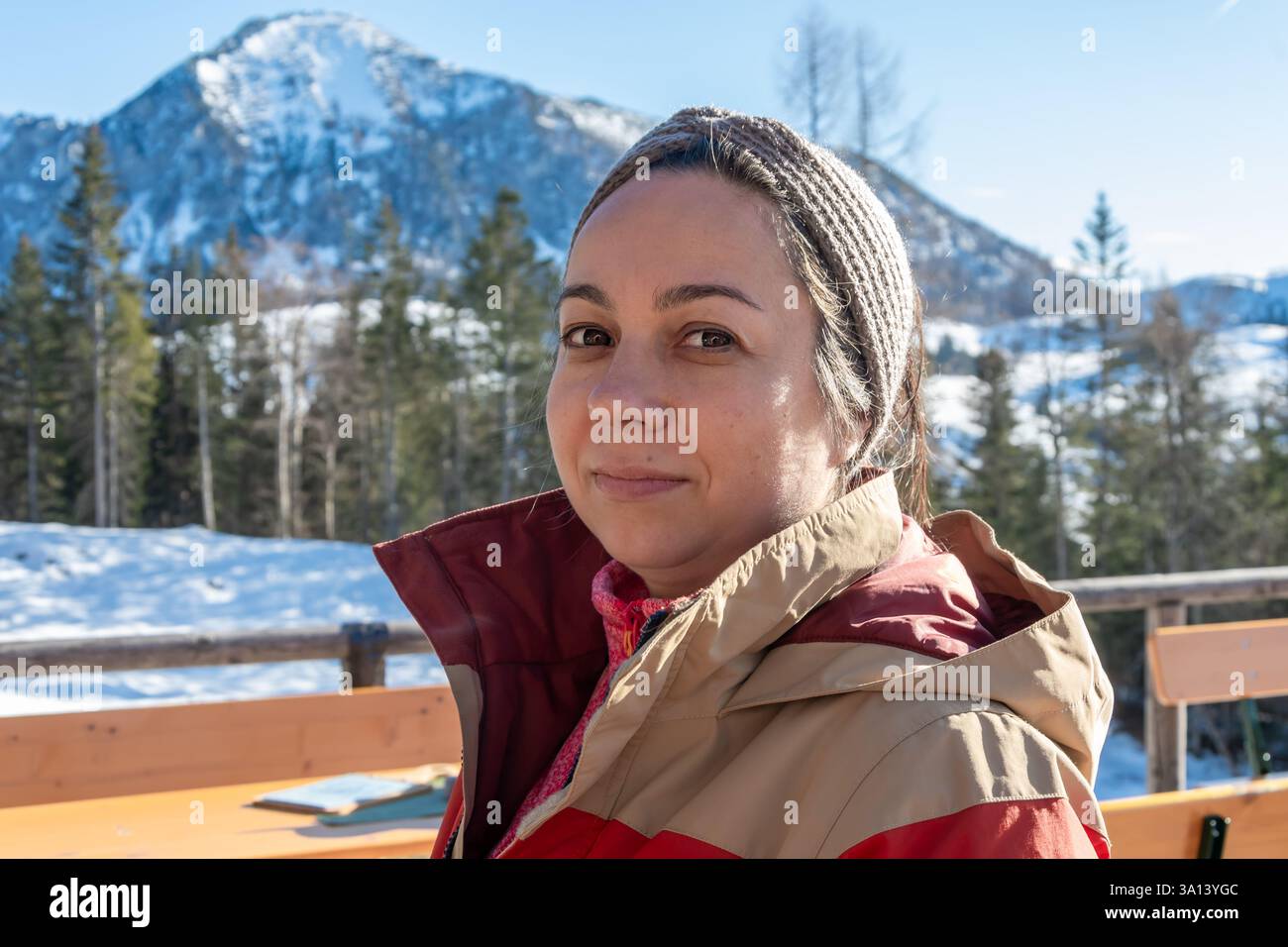 Eine 45-50-jährige Frau in warmen Winterkleidung, die auf einer Außenterrasse mit Blick auf schneebedeckte Berge und Wälder sitzt. Klares, sonniges Wetter schafft Stockfoto