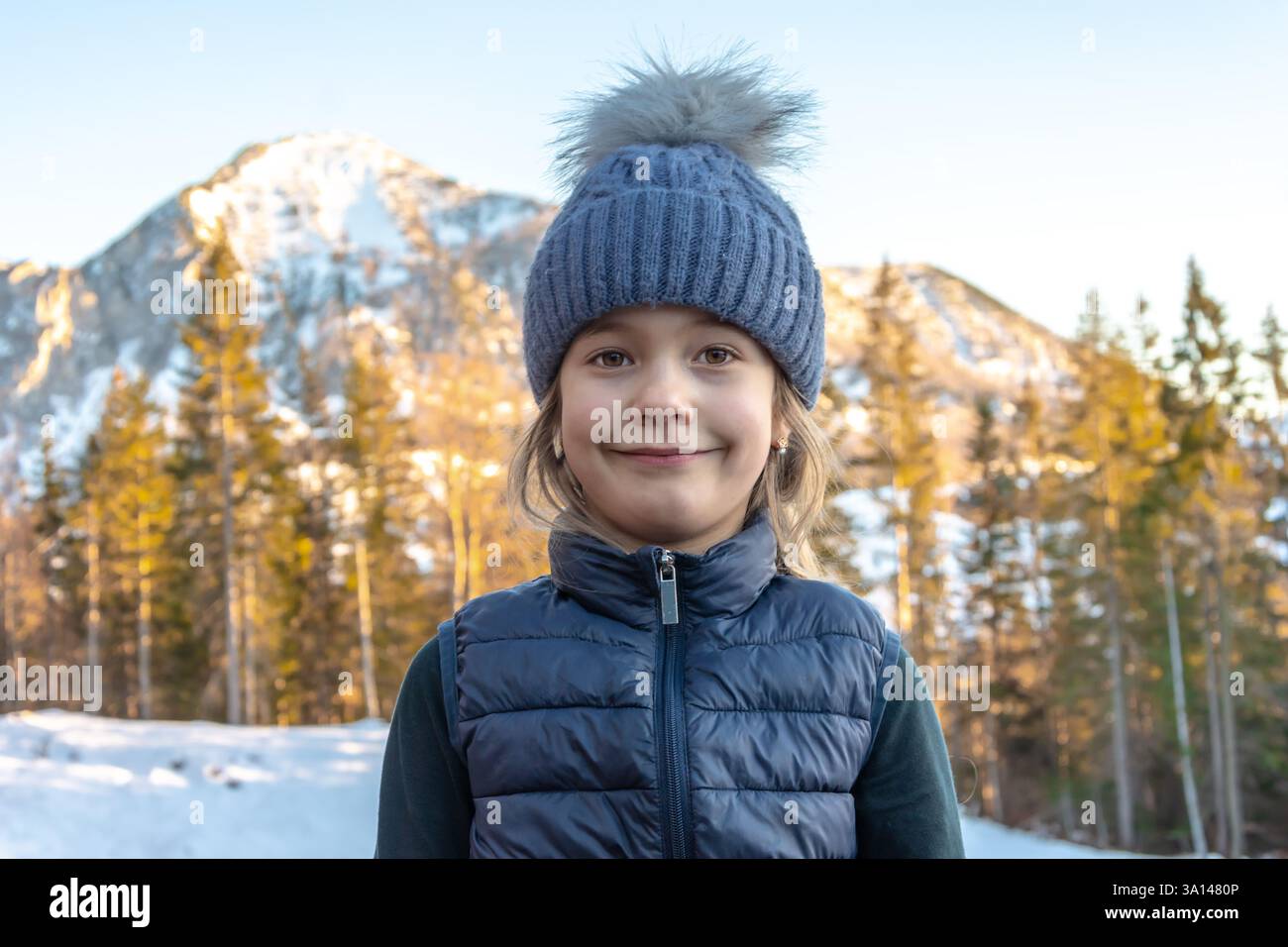 Ein Mädchen von 6-7 Jahren in einem warmen Hut mit Bommel und Daunenweste lächelt vor dem Hintergrund der schneebedeckten Berge und des Winterwaldes Stockfoto