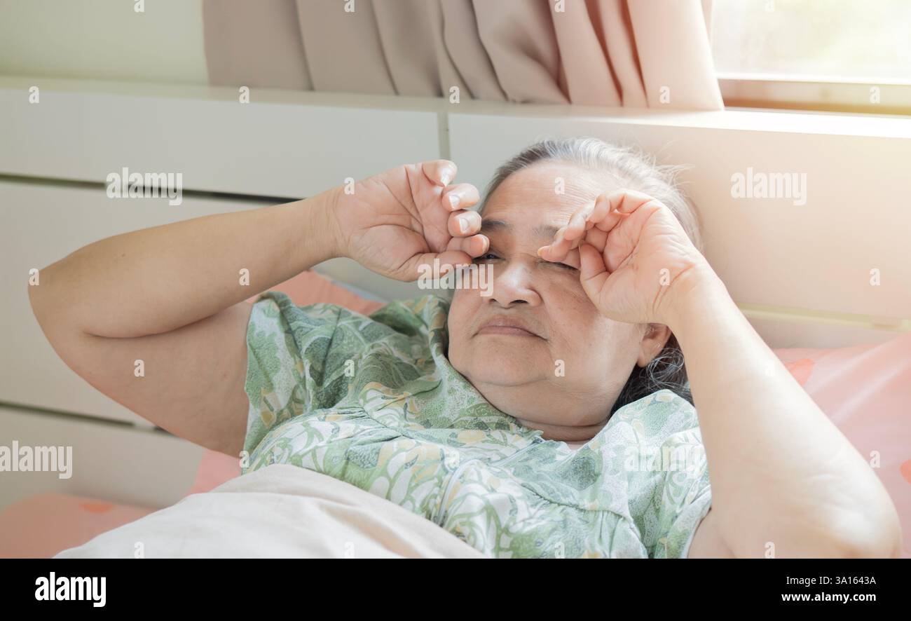 Müde, ältere asiatische Frau, die sich die Augen reibt, während die Hände im Bett liegen. Senior Person macht ein Nickerchen Stockfoto