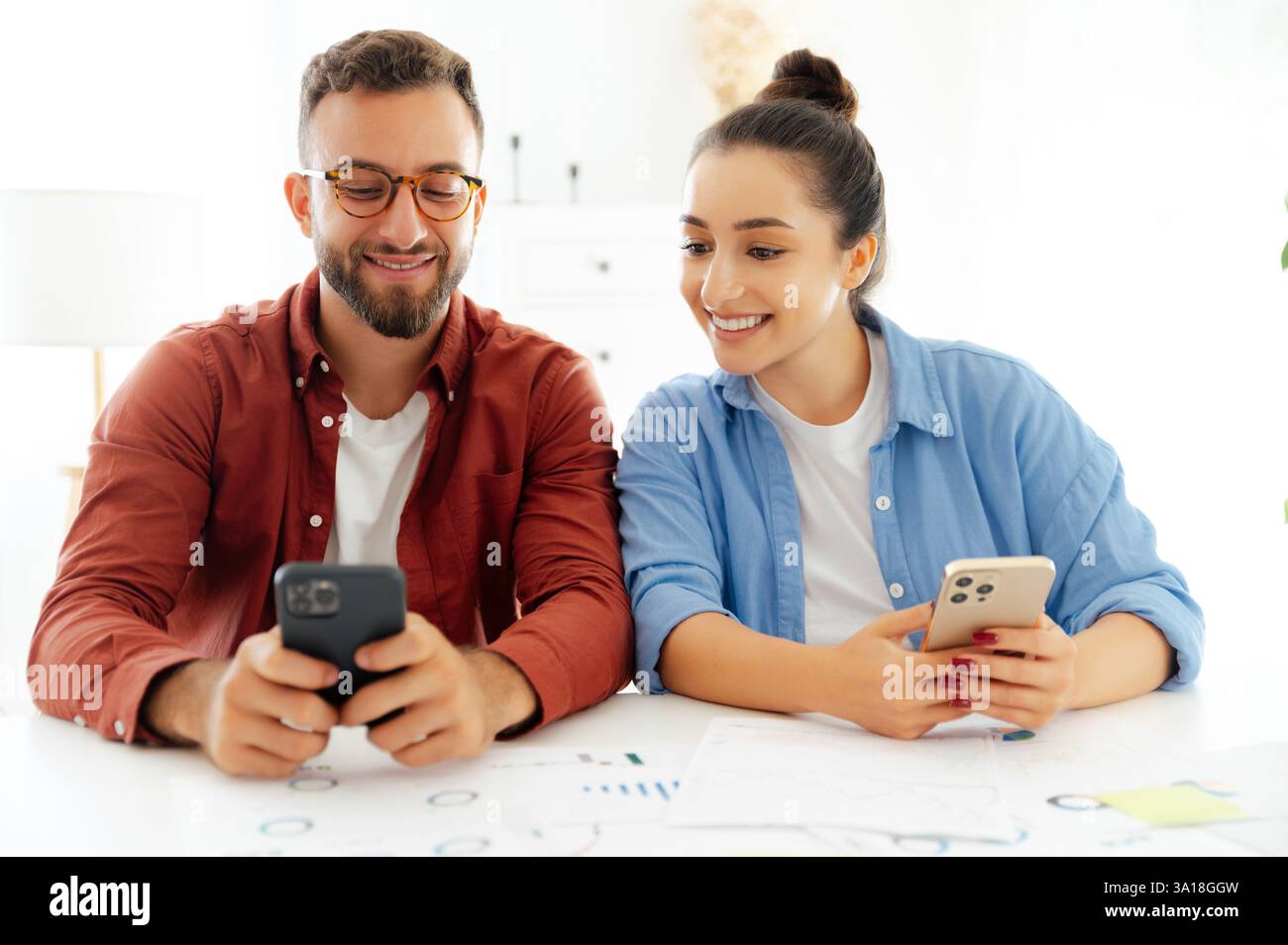 Ein Mann und eine Frau in lässiger Kleidung sitzen an einem Schreibtisch im Büro, nutzen während der Arbeitszeit Smartphones, senden online Nachrichten mit Kunden oder Freunden, surfen E-Mails, lesen Nachrichten, suchen nach Informationen Stockfoto