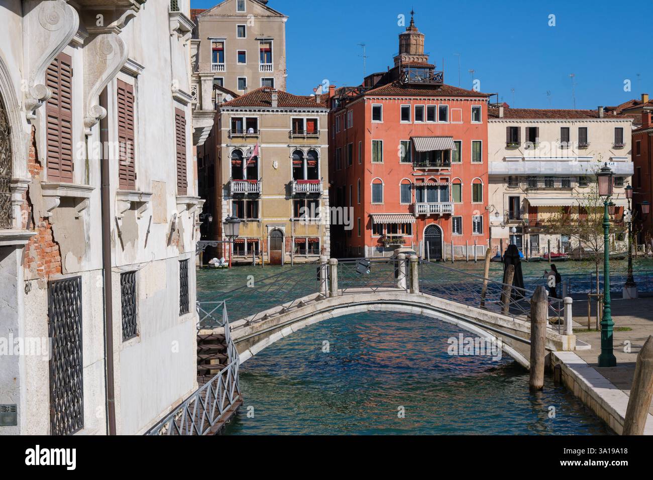 Venedig, Italien - 12. Oktober 2024: Eine kleine Fußgängerbrücke überquert einen venezianischen Kanal, umgeben von farbenfrohen historischen Gebäuden unter einem hellblauen Himmel Stockfoto